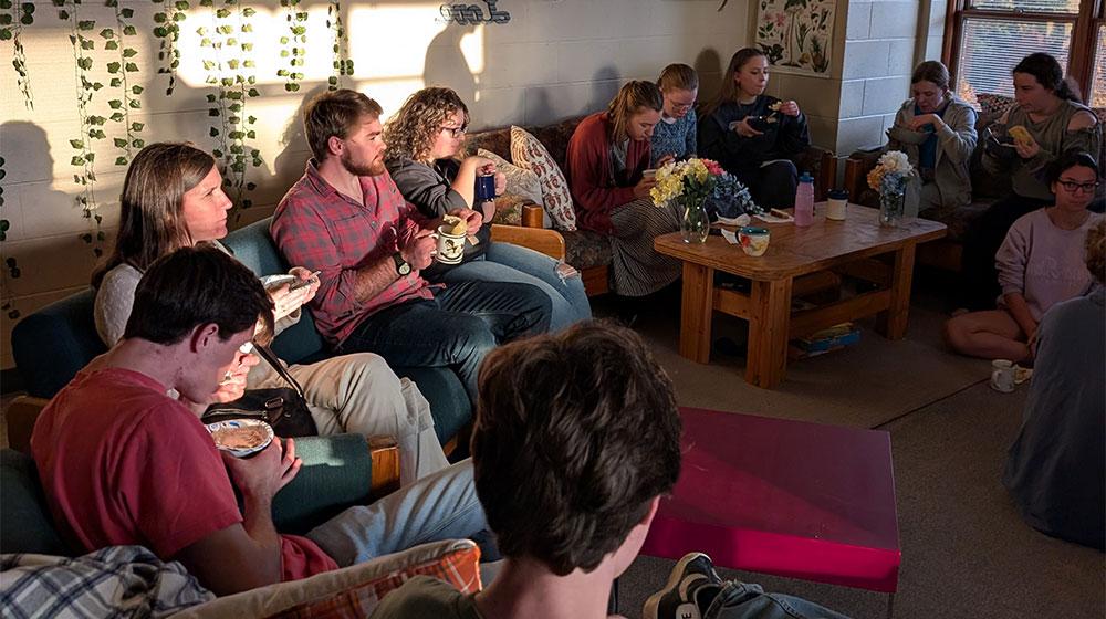 A cozy gathering of people in a warmly lit living space, enjoying food and conversation, surrounded by string lights and decorative plants.